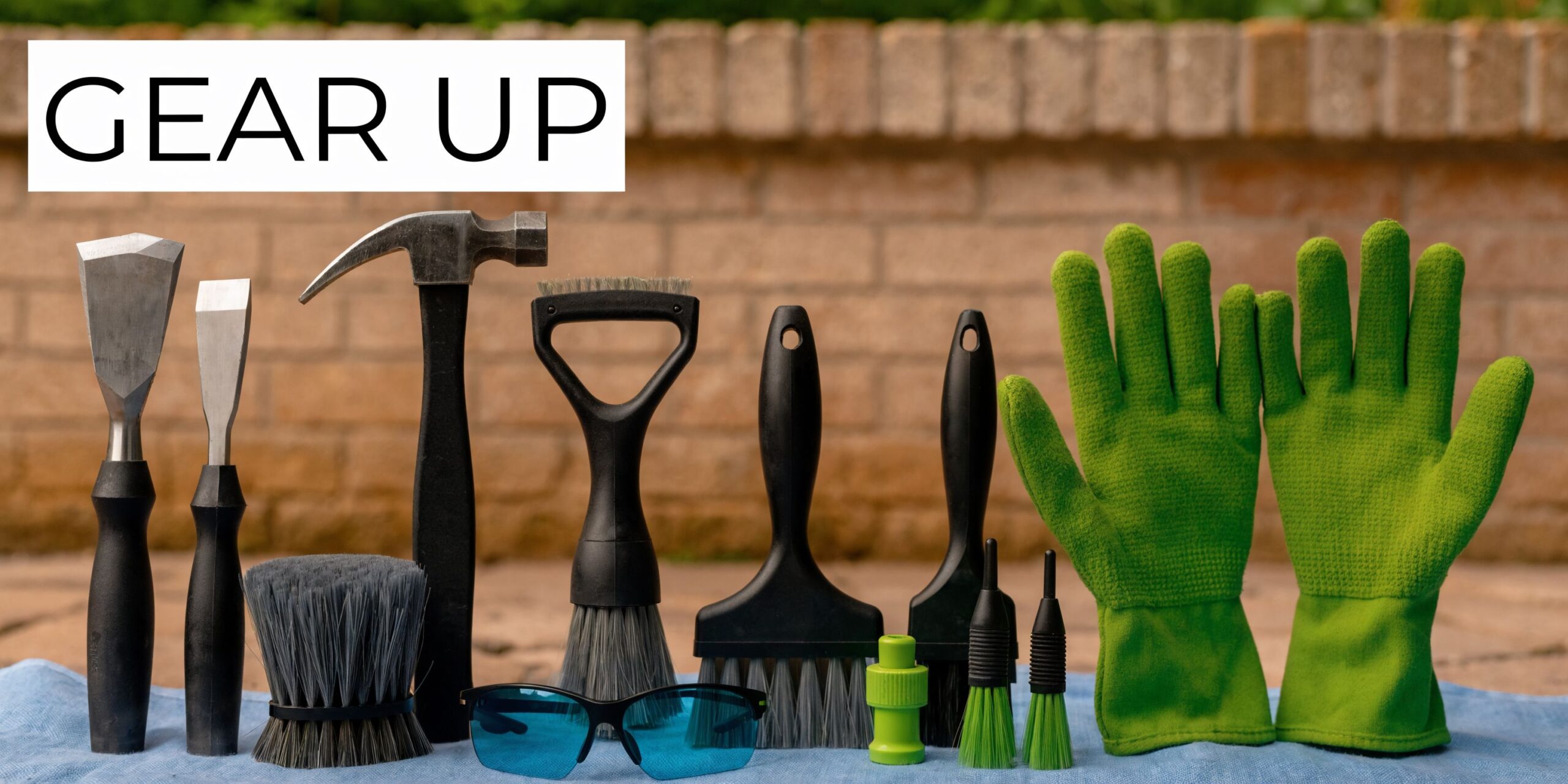 A collection of cleaning tools, brushes, safety glasses, and gardening gloves laid out against a brick wall.