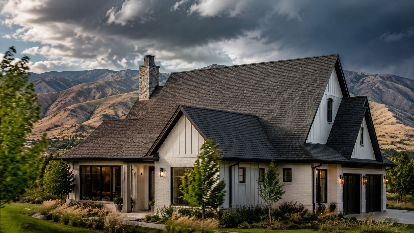 A luxurious modern farmhouse with a dark shingle roof nestled against a dramatic mountain range landscape.